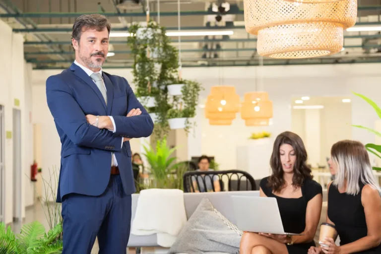 Confident businessman in blue suit standing in modern office while two women discuss work on laptop in background