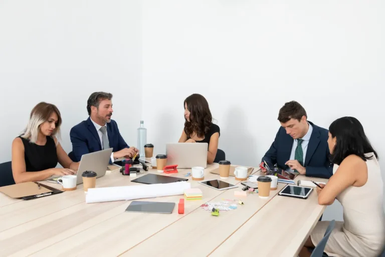 Business team in formal meeting discussing strategy with laptops, tablets, coffee cups, and office supplies on table.
