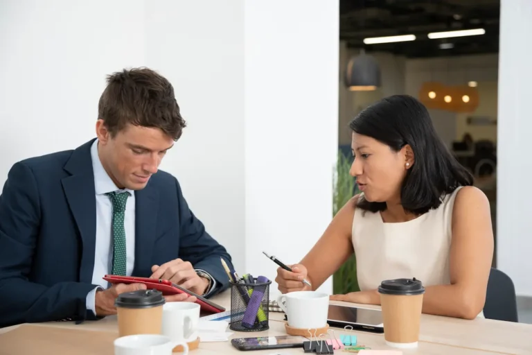 Two business professionals discussing work at a modern office table with tablets, coffee, and stationery.